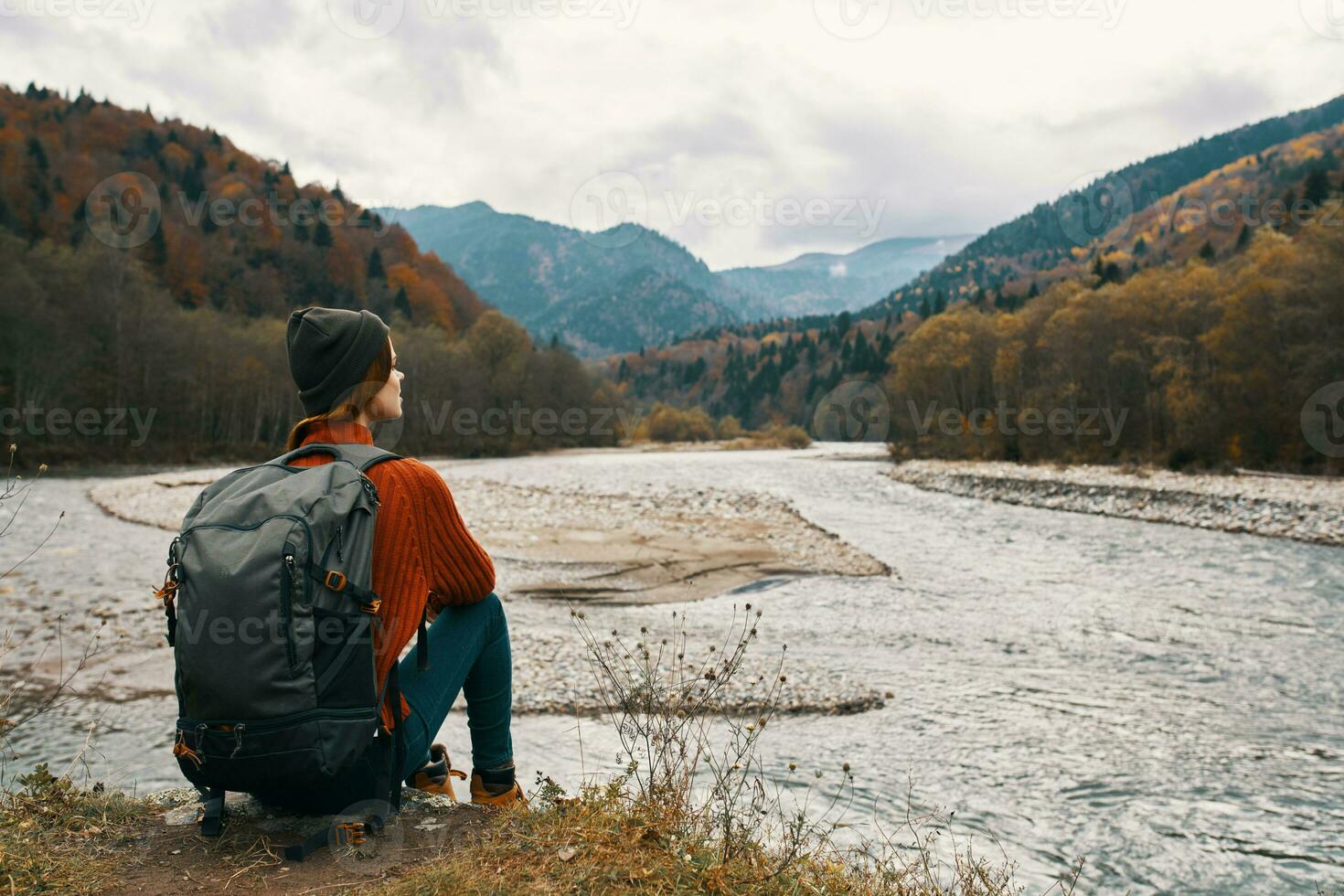woman with a backpack in a jacket and a hat on the river bank in the mountains side view photo