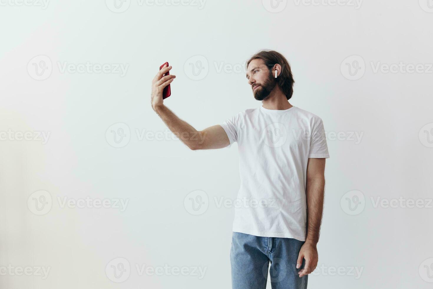 A male blogger videotaping himself on his phone and chatting with people online with a smile in a white T-shirt against a white wall photo