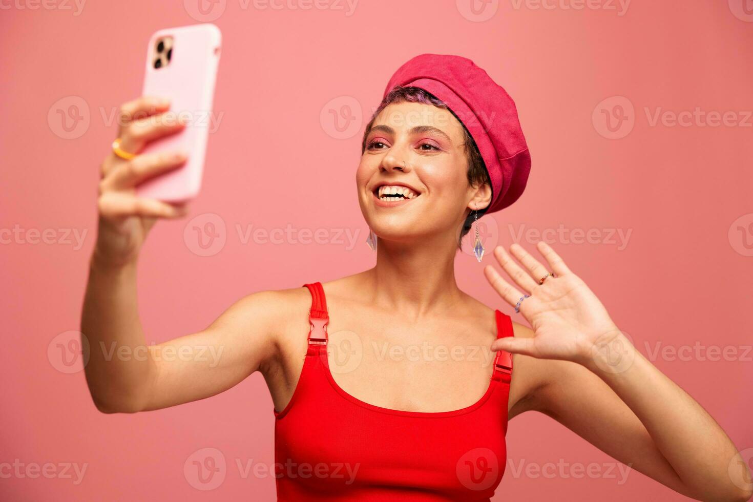 A young woman blogger with colored pink hair and a short haircut takes a picture of herself on the phone and broadcasts a smile in stylish clothes and a hat on a pink background monochrome style photo