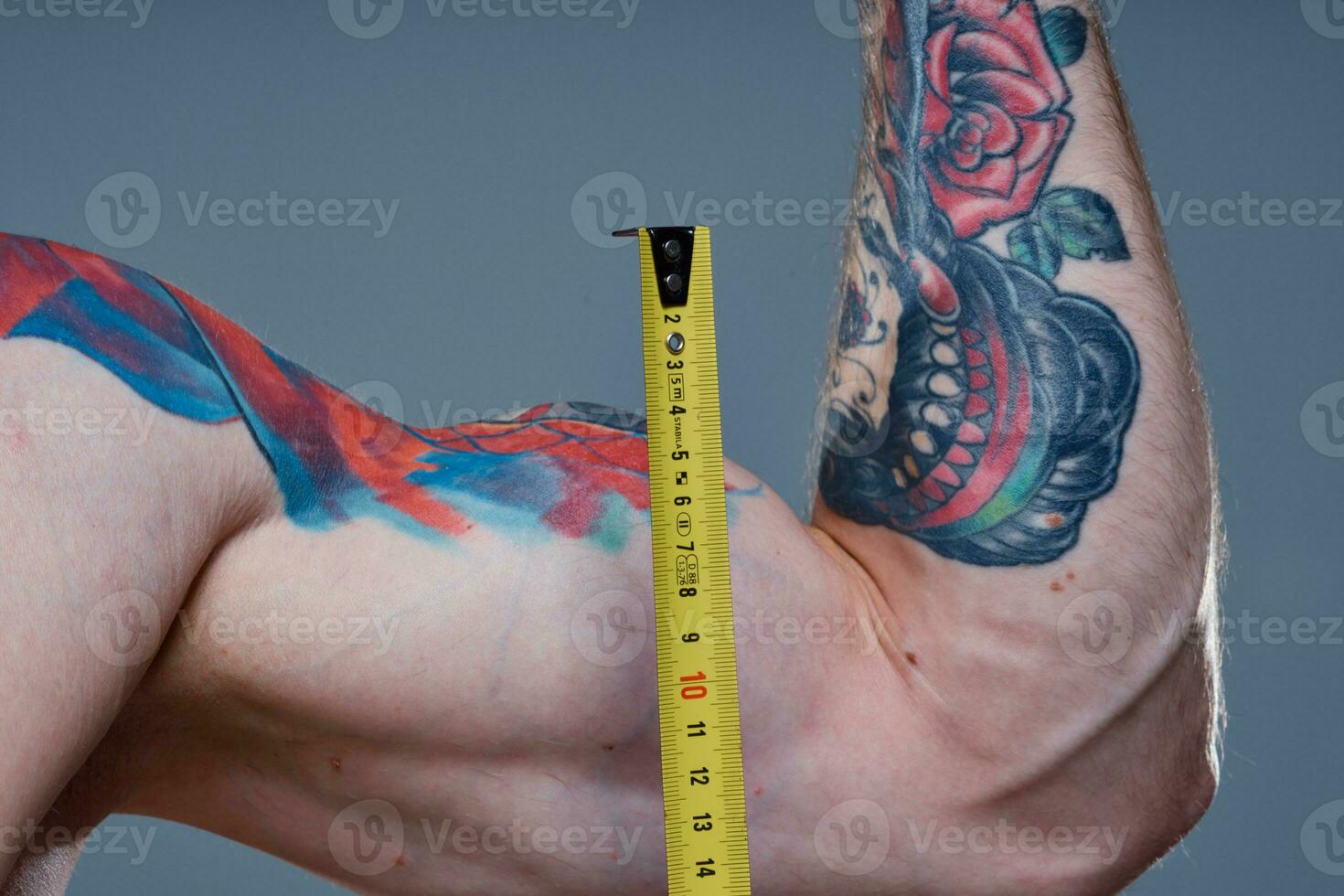 guy measures arm muscles with centimeters on a gray background and a multicolored tattoo bodybuilder fitness photo