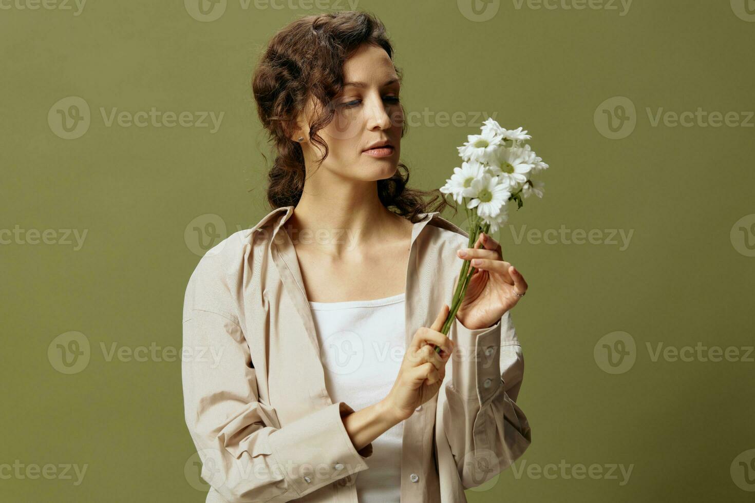 Calm cute cheerful curly beautiful female in linen casual shirt hold chamomiles looks with love at flowers posing isolated on over olive green pastel background. Nature is beauty concept. Copy space photo