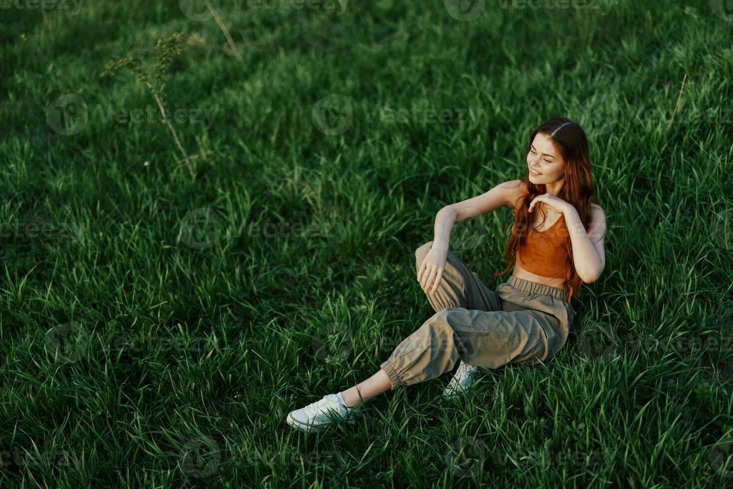 A beautiful woman with long, curly red hair sits in the park in the summer on the green grass in the sunset light photo