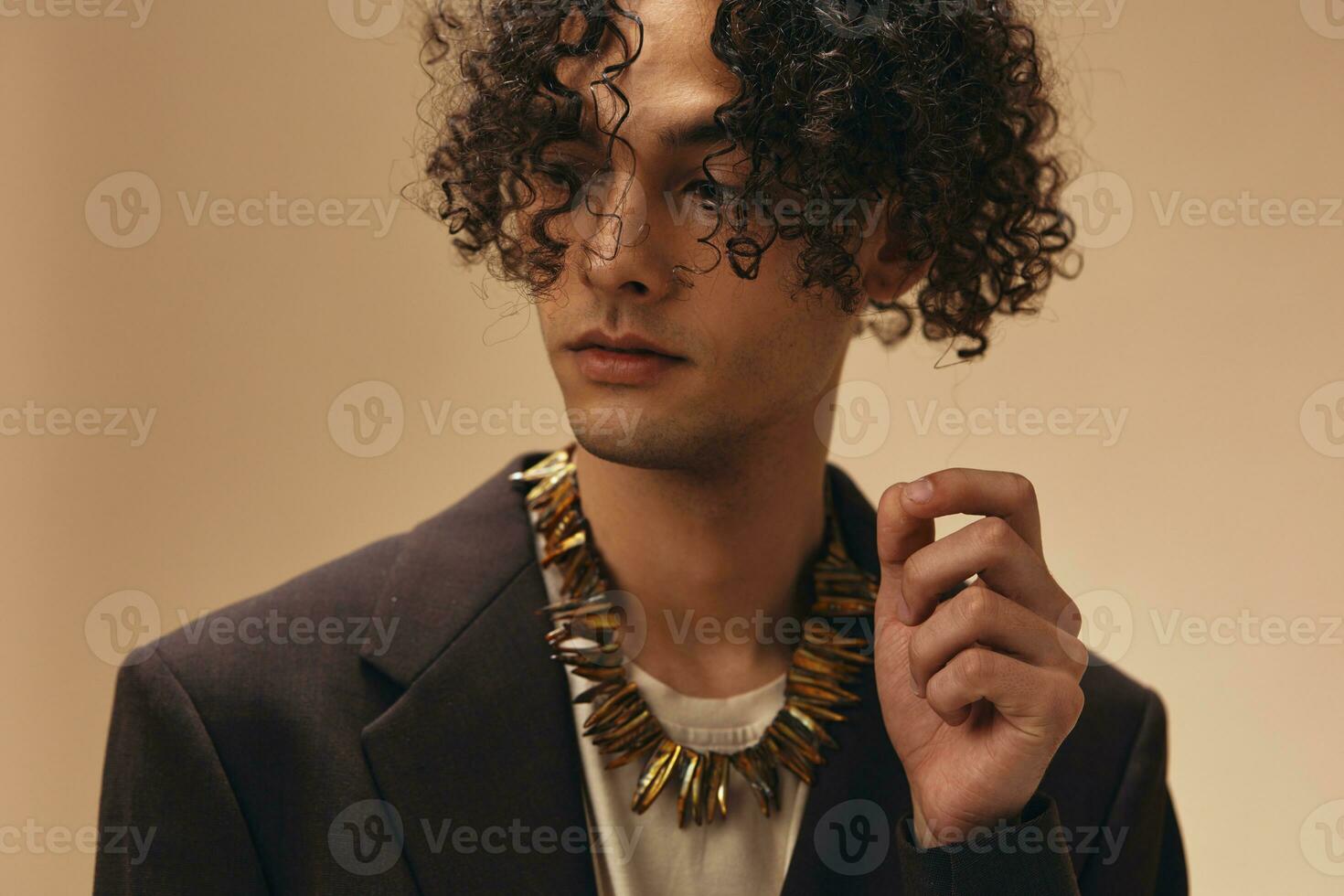 Awesome tanned curly man in classic brown jacket touches ethnic necklace posing isolated on over beige pastel background. Fashion New Collection offer. Retro style concept. Free place for ad photo