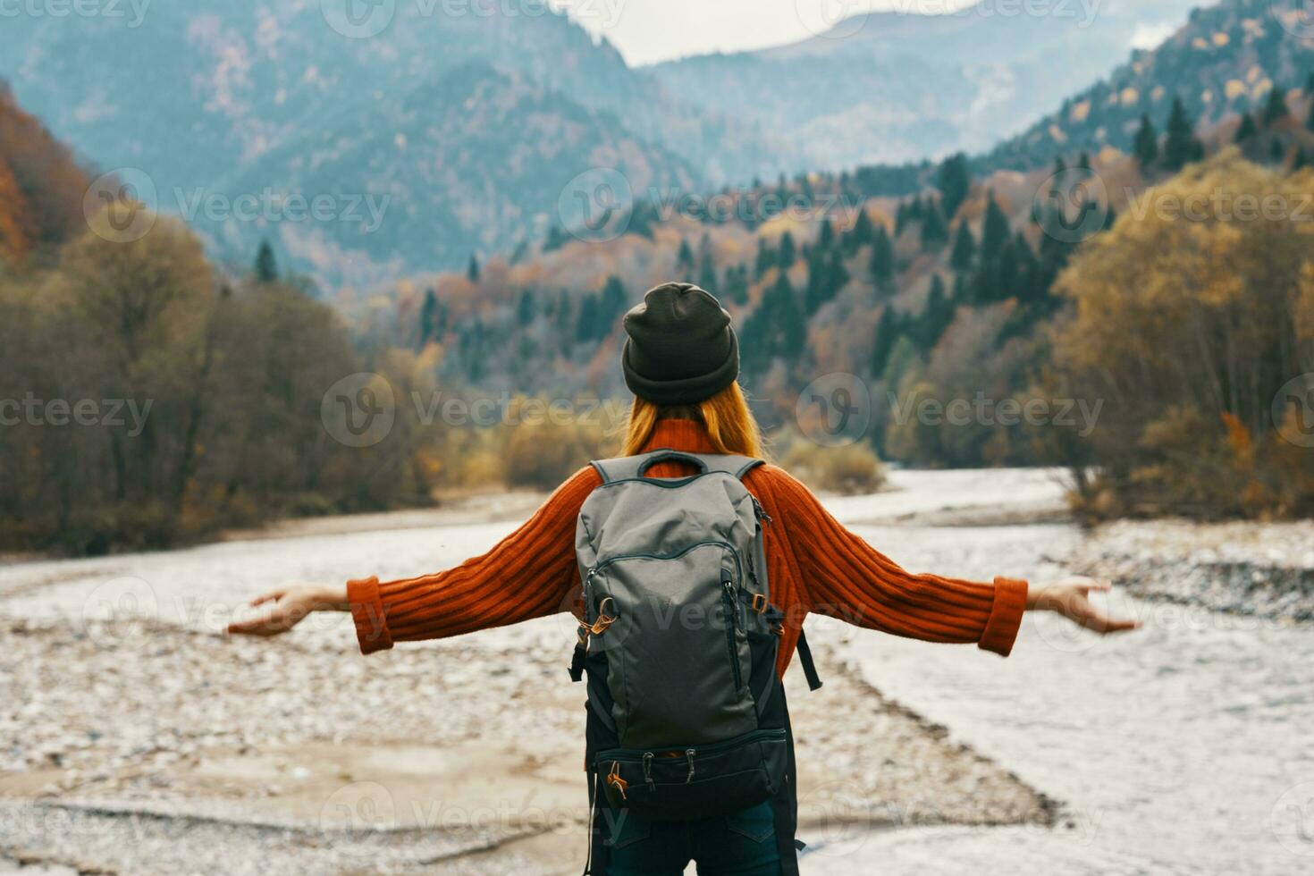 joyful woman tourist with a backpack looks at the mountains near the river on the bank photo