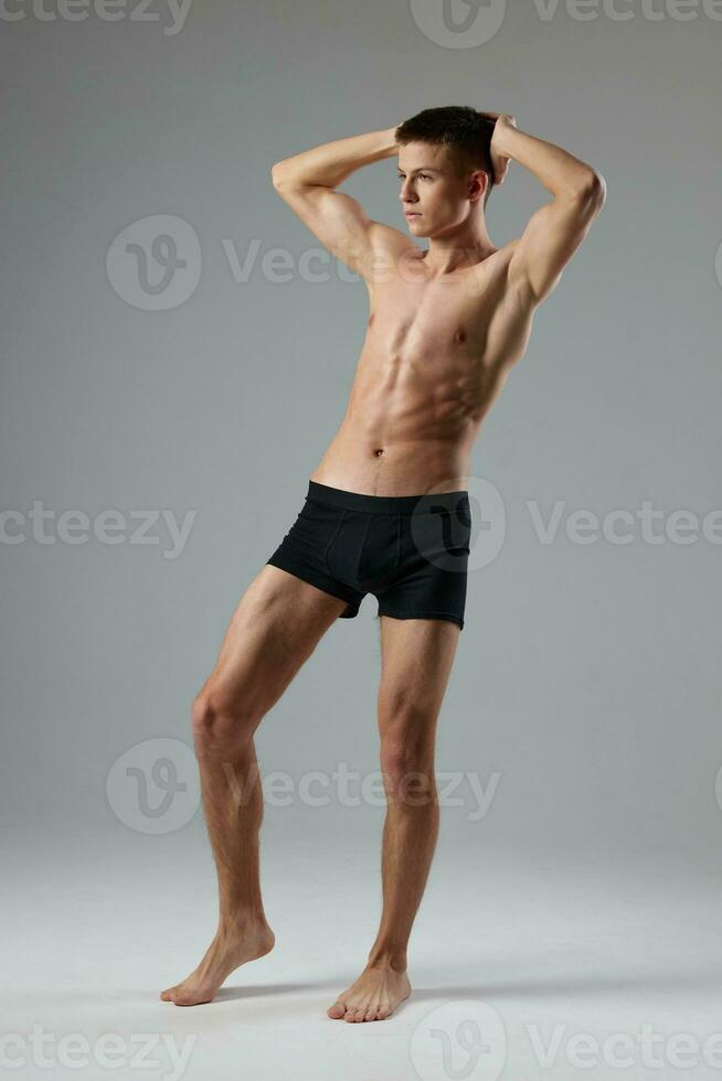 athlete in black shorts holds hands behind his head naked torso gray room full-length model photo