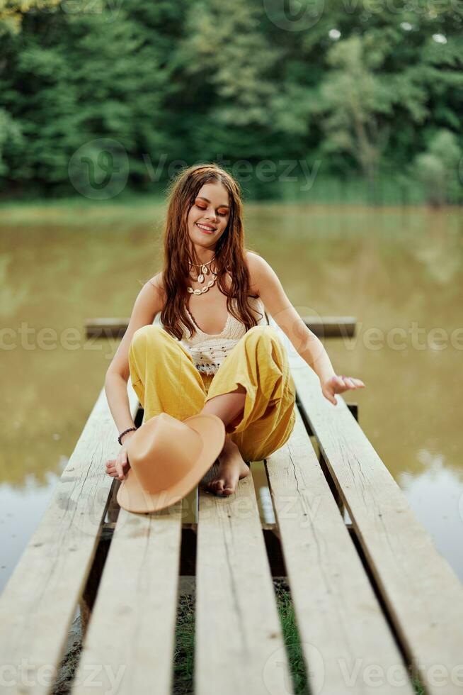 A hippie woman sits with her hat on a bridge by a lake with her hands up in the air on a nature trip and smiling happily in eco-clothing. Relaxed lifestyle photo