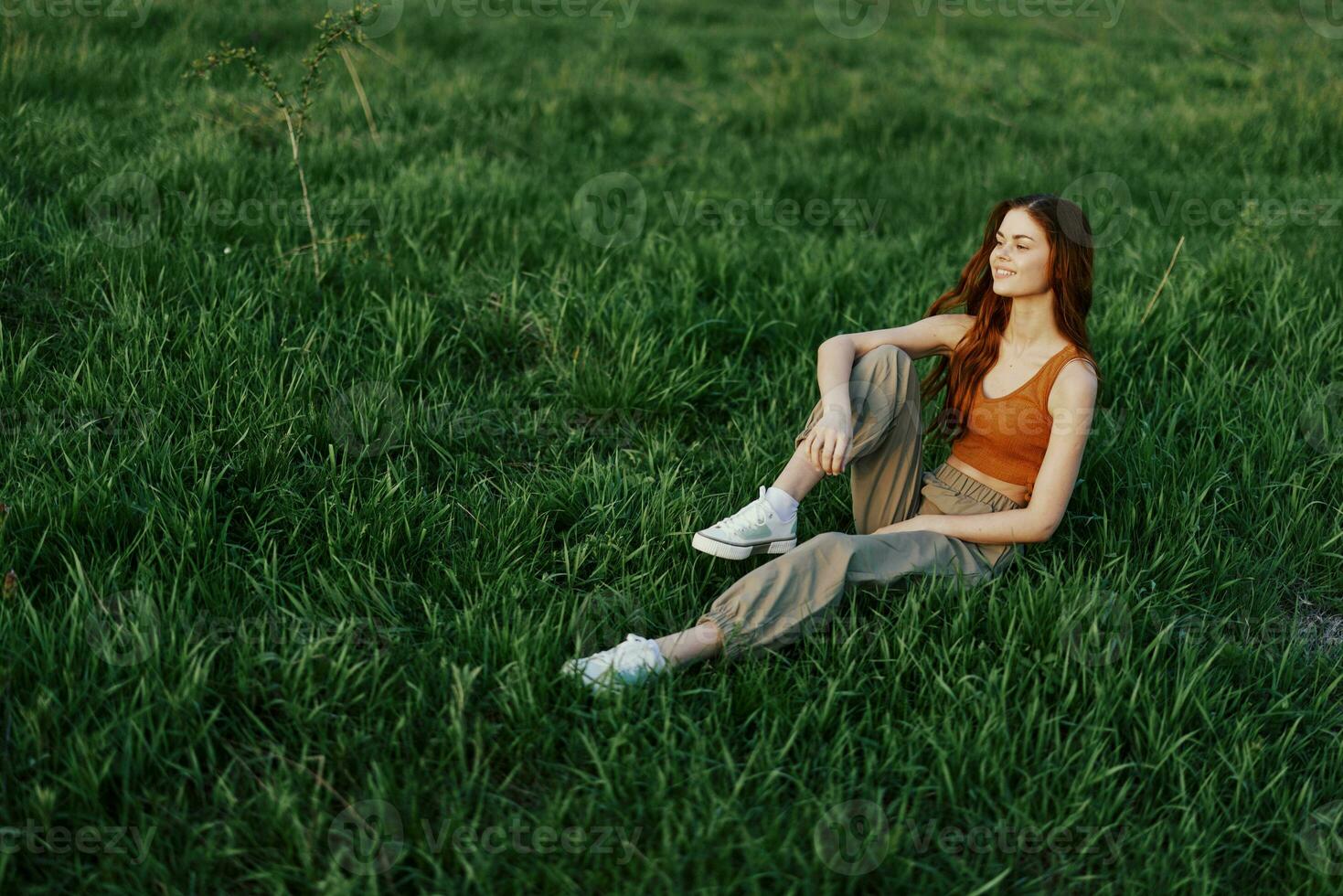 The redheaded woman sits in the park on the green grass wearing an orange top, green pants, and sneakers and looks out at the setting summer sun photo