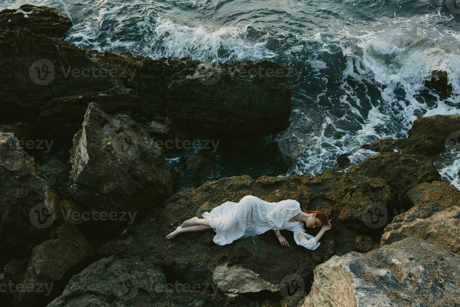 beautiful young woman in long white dress wet hair lying on a rocky cliff landscape photo