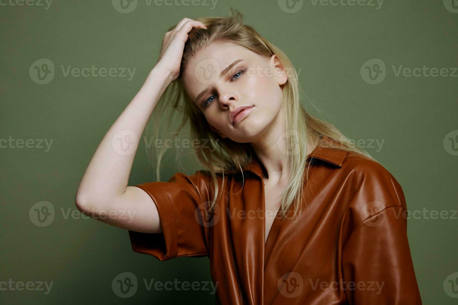 Fashionable stylish woman touching hair posing isolated in brown palette leather dress trench coat isolated on over olive green wall background. Cool offer for brands photo