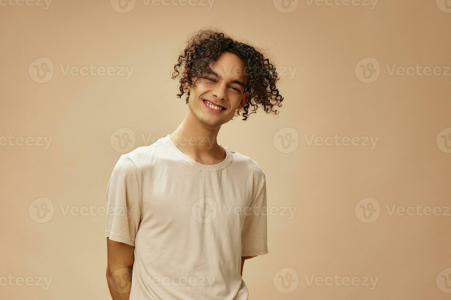 Cheerful cute awesome tanned curly man in basic t-shirt posing isolated on over beige pastel background. Fashion New Collection offer. People and Emotions concept. Free place for ad photo