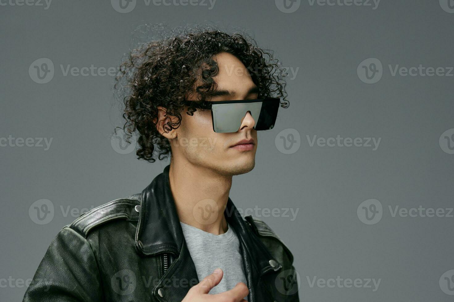 Stylish tanned curly man leather jacket mirror trendy eyewear looks aside posing isolated on gray studio background. Cool fashion offer. Huge Seasonal Sale New Collection concept. Copy space for ad photo