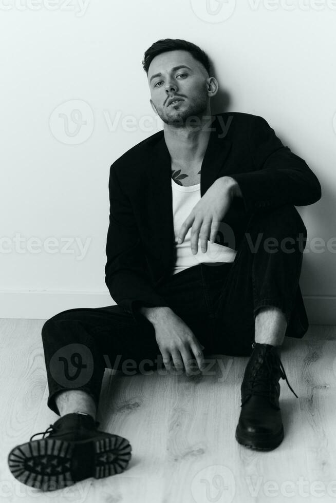 Modelling snapshots. Serious self-confident attractive handsome man in black jacket sitting on floor posing in white studio background. Black and White concept. Copy space photo