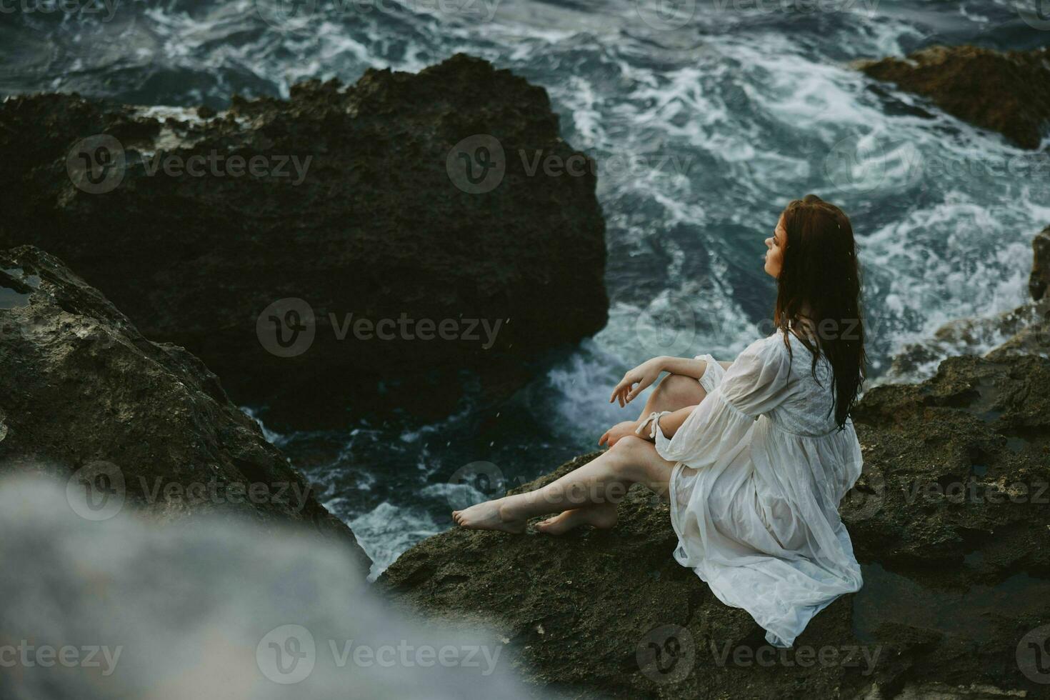a woman with wet hair in a white dress sits on a cliff barefoot unaltered photo