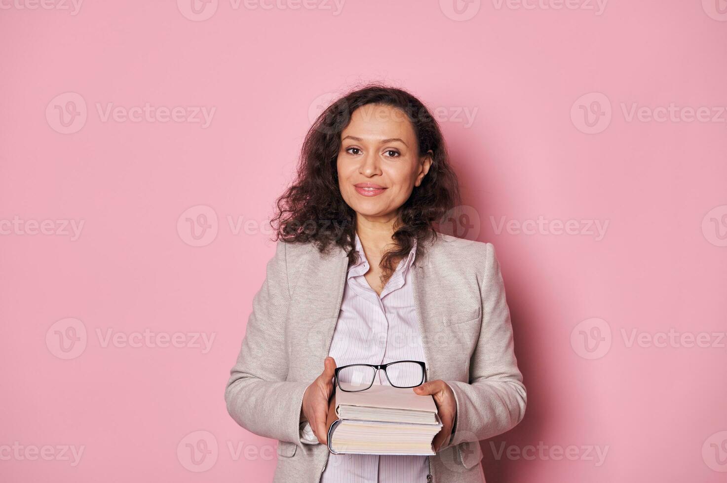 Attractive school teacher librarian smiling at camera, holding book on pink background. Education. Erudition. Knowledge photo