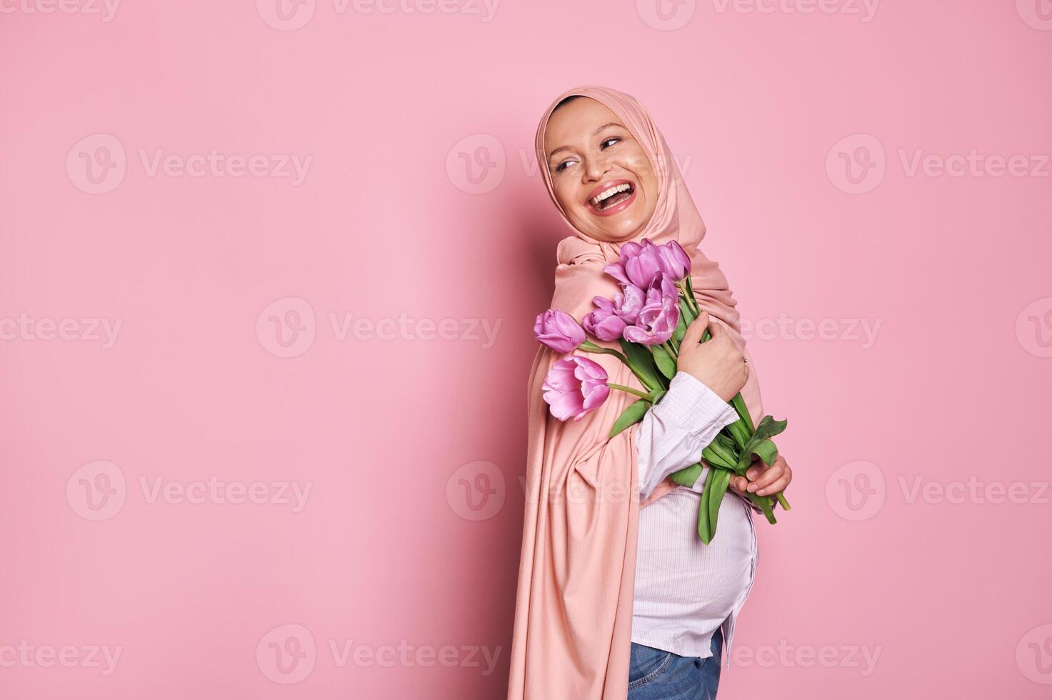 Pregnant Arab Muslim woman in hijab holds a bouquet of tulips for Mother's Day, smiles looking aside, pink background photo