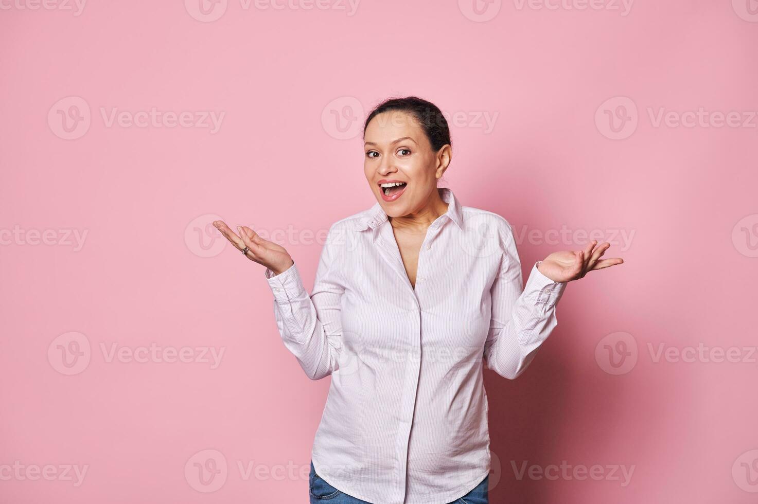 encantador asombrado embarazada mujer participación su manos palmas arriba, expresando asombro y sorpresa en rosado antecedentes foto