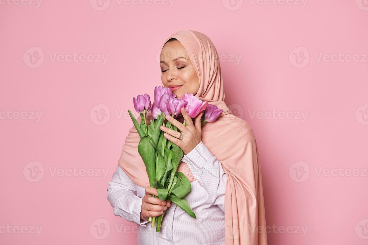 encantador musulmán mujer en rosado hiyab, olfateando ramo de flores de tulipanes, posando con su ojos cerrado en aislado rosado antecedentes foto