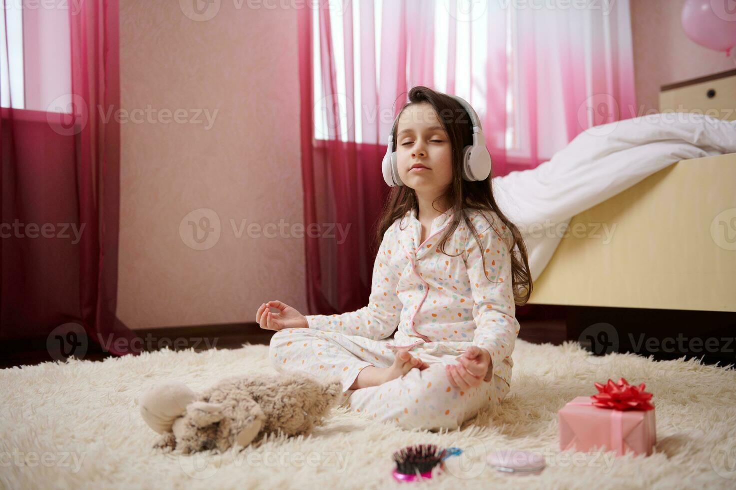 Lovely baby girl in wireless headphones, wearing stylish pajamas, sitting on the carpet and meditating in lotus position photo