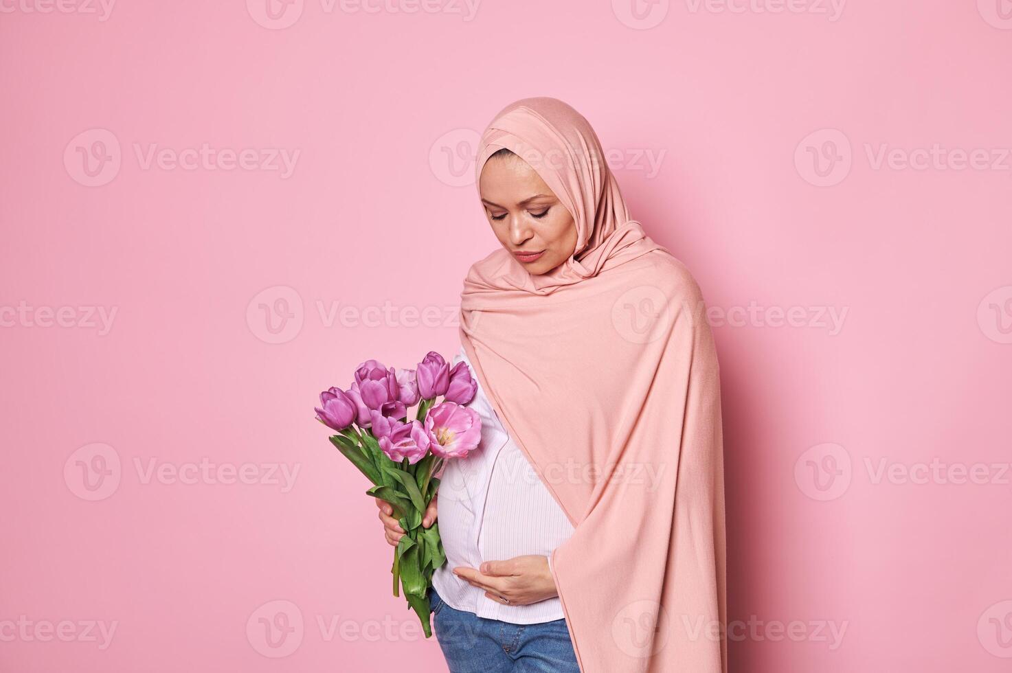 Beautiful Muslim woman with a bouquet of tulips, touching her pregnant belly, isolated on pink background. Copy space photo