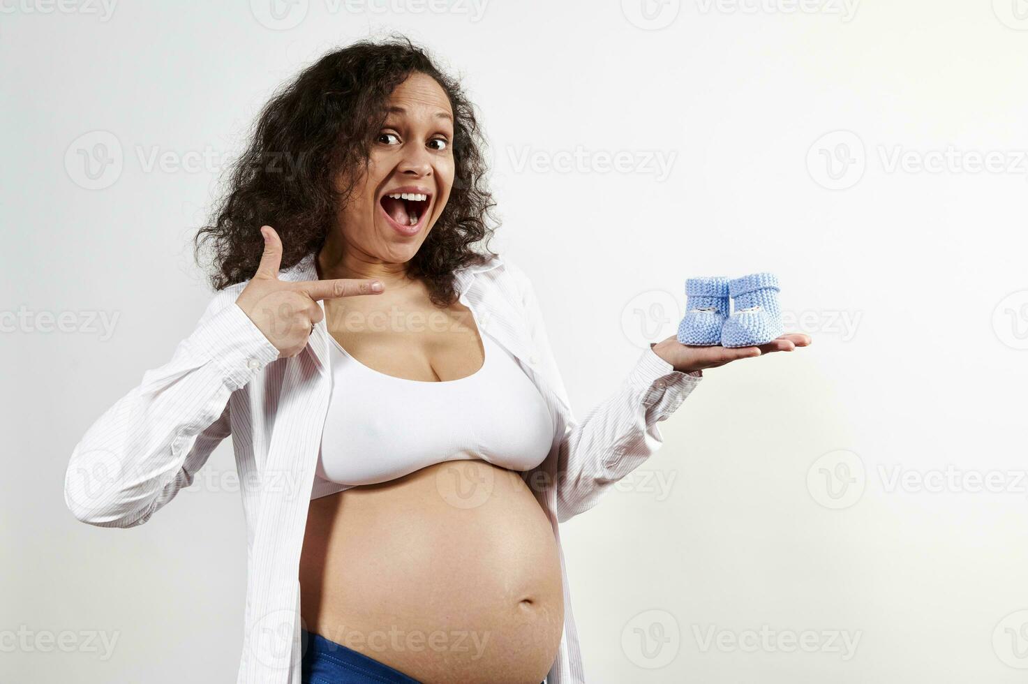 Excited pregnant woman smiles broadly looking at camera, points at blue baby booties in her hand, on white background photo