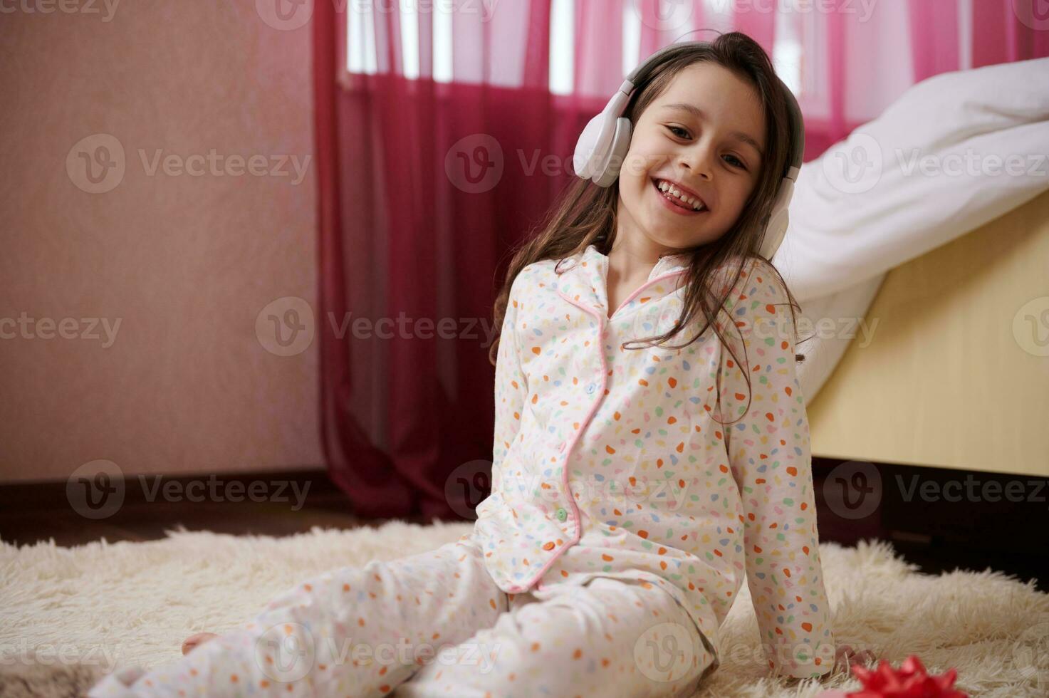 Little child girl in wireless headphones, sitting on a carpet, dressed in stylish pajamas, listening to soothing music photo