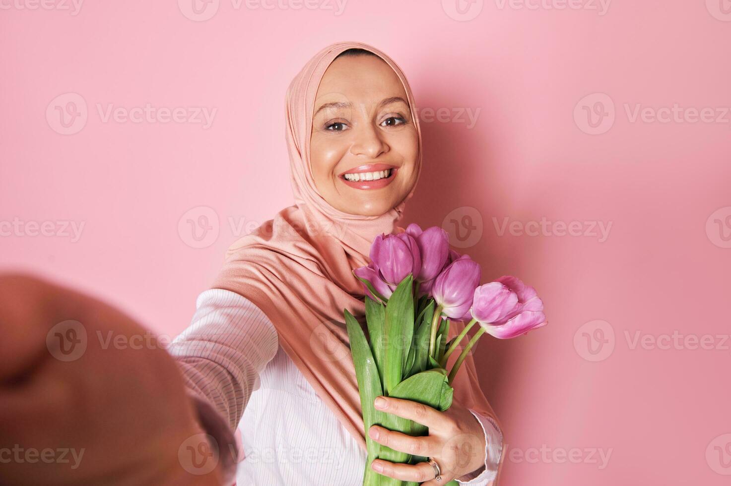 sonriente musulmán mujer en rosado hiyab, hace un autofoto, posando con un ramo de flores de púrpura tulipanes en aislado rosado antecedentes foto