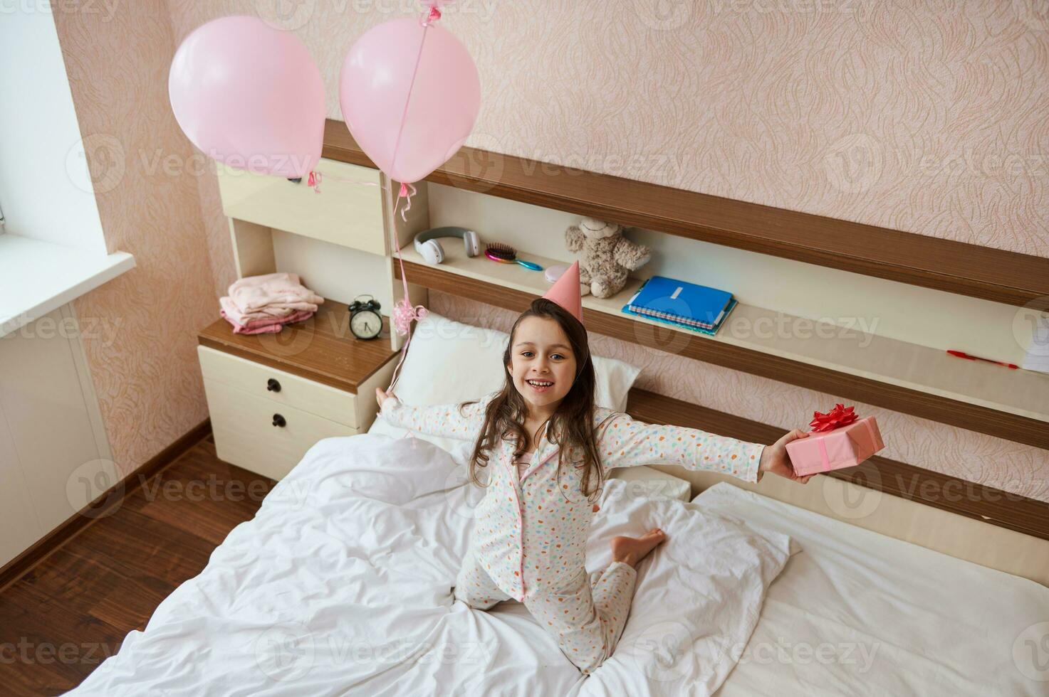 Top view birthday girl in party hat, holding bunch of pink balloons and gift box, sitting on bed and smiling into camera photo