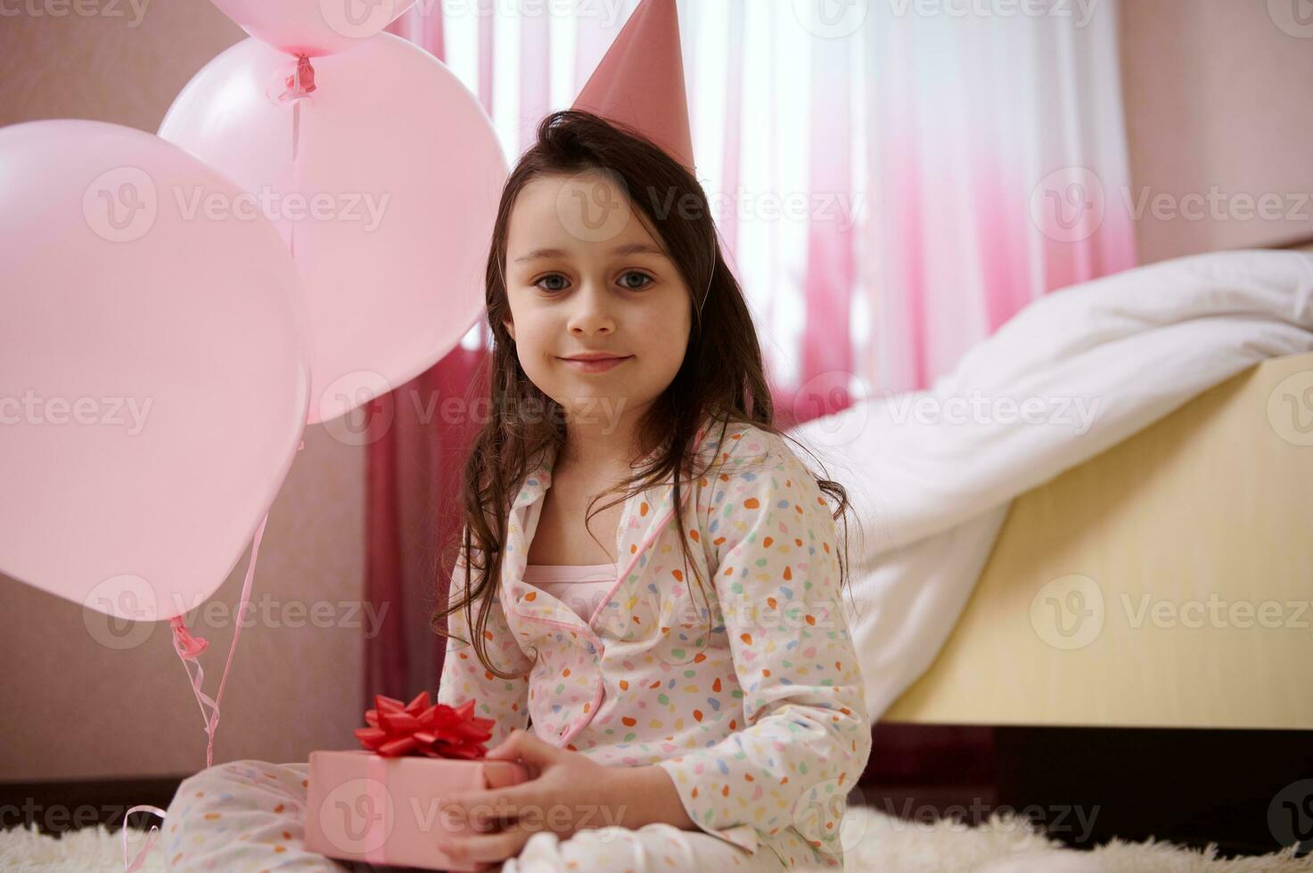 contento pequeño niño niña en pijama y rosado fiesta sombrero, sostiene un linda regalo caja para cumpleaños y sonrisas mirando a cámara foto