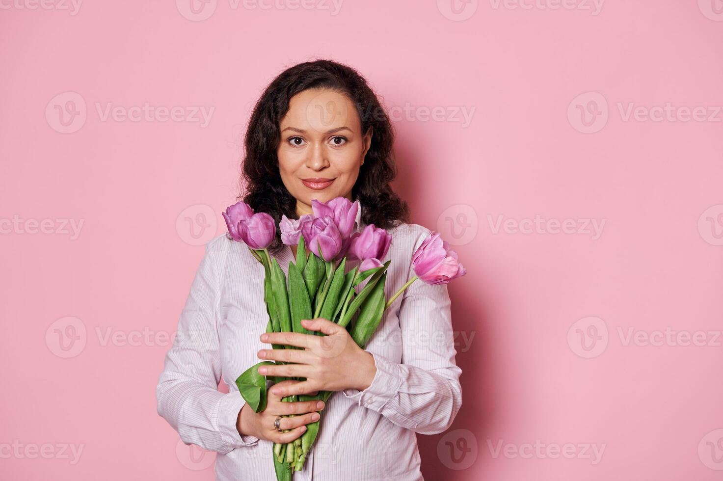 Confident beautiful woman with a bouquet of purple tulips, smiling looking at camera over isolated pink background photo