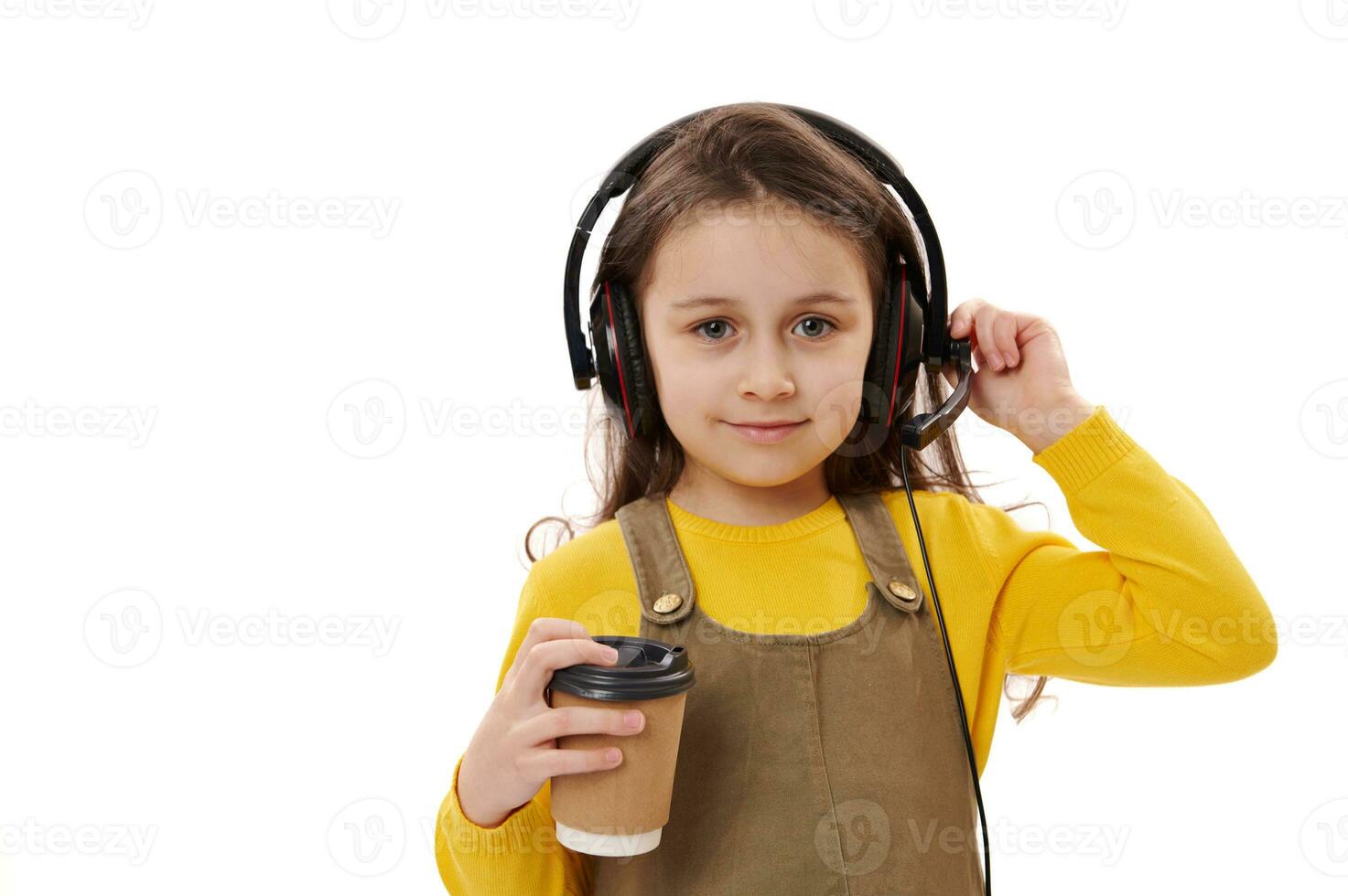 Beautiful Caucasian little girl wearing headphones, holding disposable paper cup with hot drink, smiling at camera photo