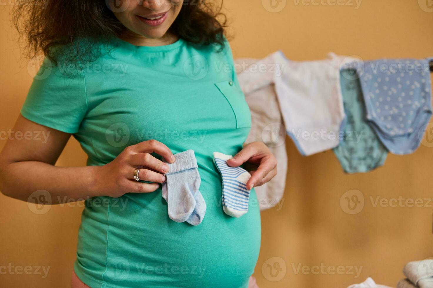 Closeup of pregnant woman holding blue baby socks on her belly, standing against ironed bodysuits for her newborn child photo