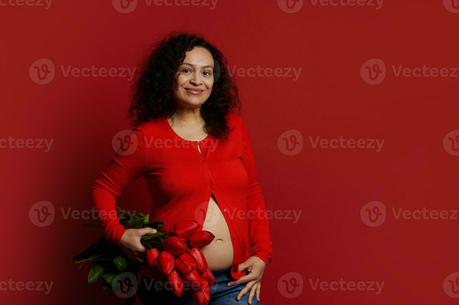 Multi ethnic pregnant woman smiles looking at camera, holds a bouquet of red tulips on isolated color background photo