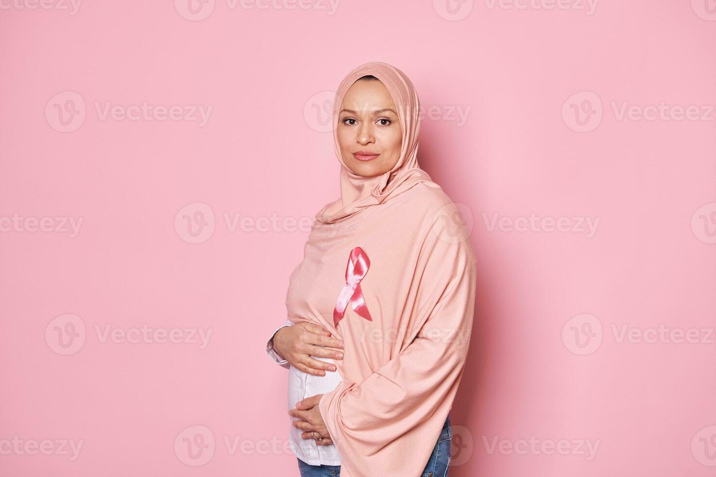 confidente joven Medio este musulmán embarazada mujer en rosado hiyab, vistiendo un satín conciencia cinta, aislado en rosado foto