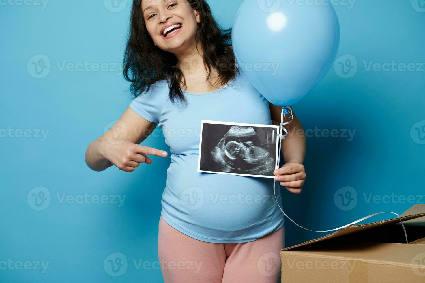 Happy cheery gravid woman smiling at camera, poiting at ultrasound image of her newborn baby, isolated blue background photo