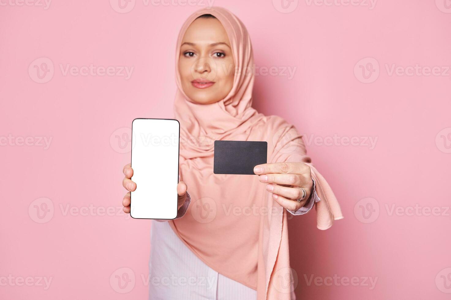 detalle en negro el plastico banco crédito tarjeta y teléfono inteligente con blanco vacío pantalla en manos de musulmán mujer en rosado hijab foto