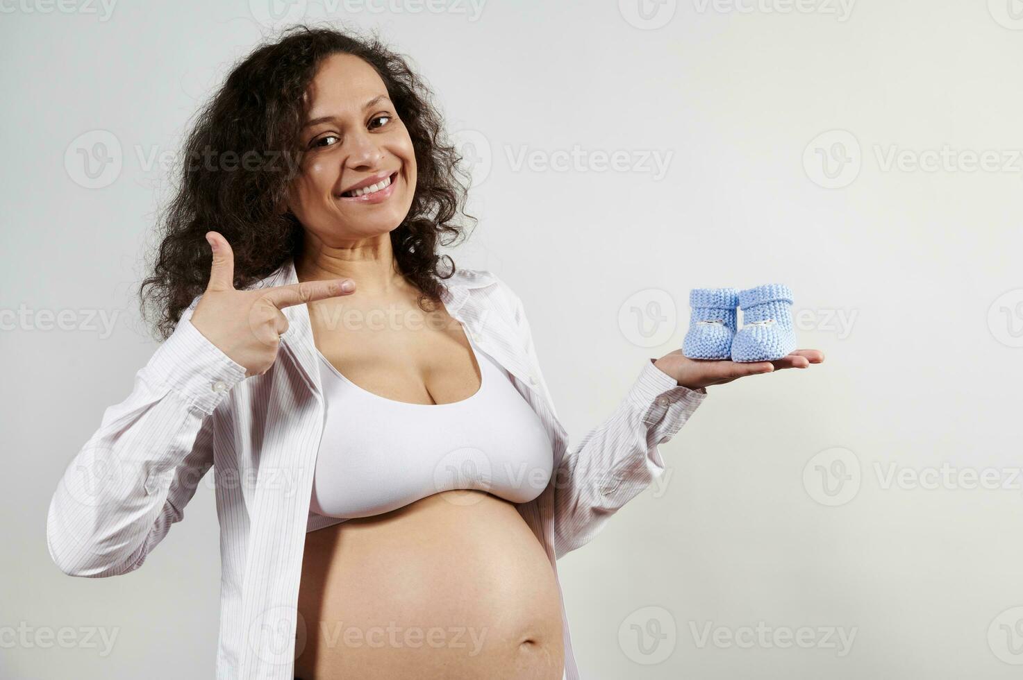 Attractive delightful smiling pregnant woman pointing finger at blue knitted baby booties, isolated on white background photo