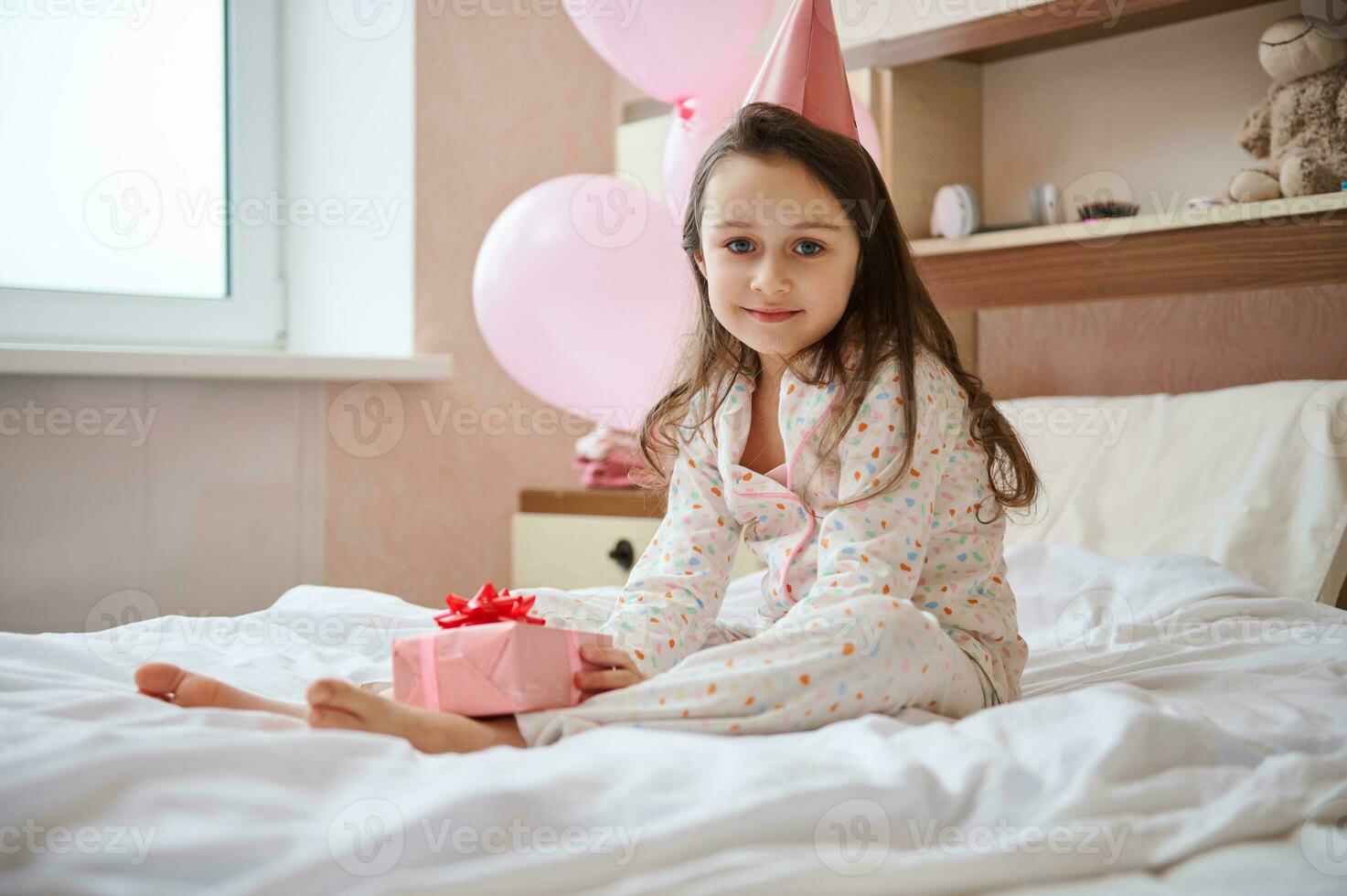 Lovely birthday girl in pink festive hat, holding gift box, sitting on the bed in her bedroom with helium balloon photo