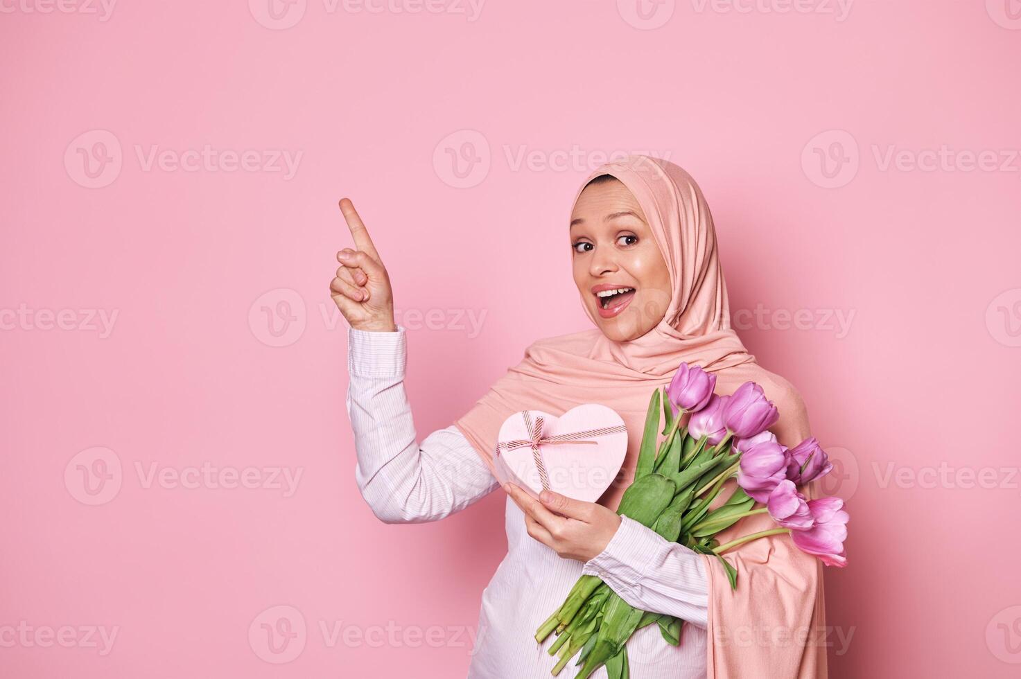 Delighted pregnant Muslim woman with a bouquet of tulips and a cute present, pointing at copy space on pink background photo