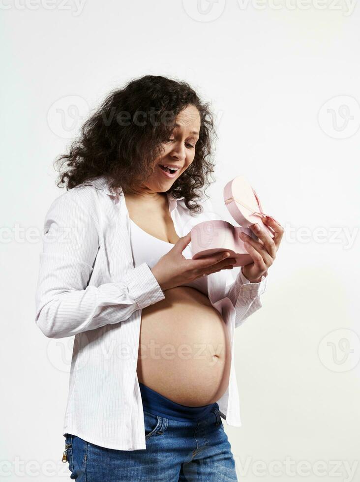 Multi ethnic pregnant woman, expressing positive emotions opening gift box for special occasion, isolated on white photo