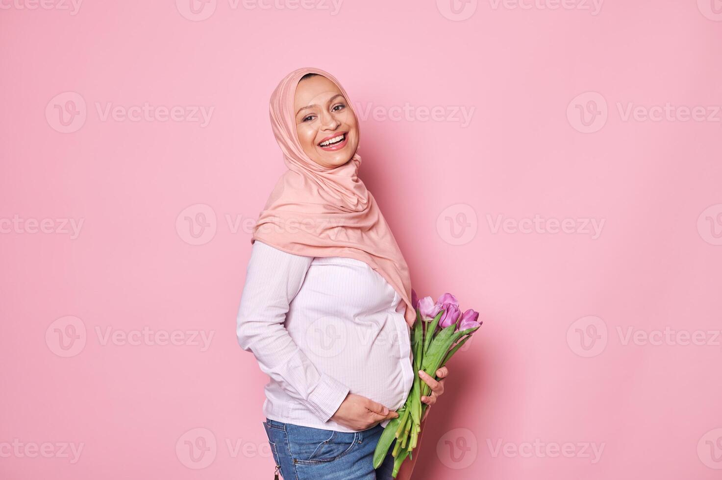 Happy Muslim pregnant woman in pink hijab, smiling with beautiful smile, touching her tummy, isolated on pink background photo