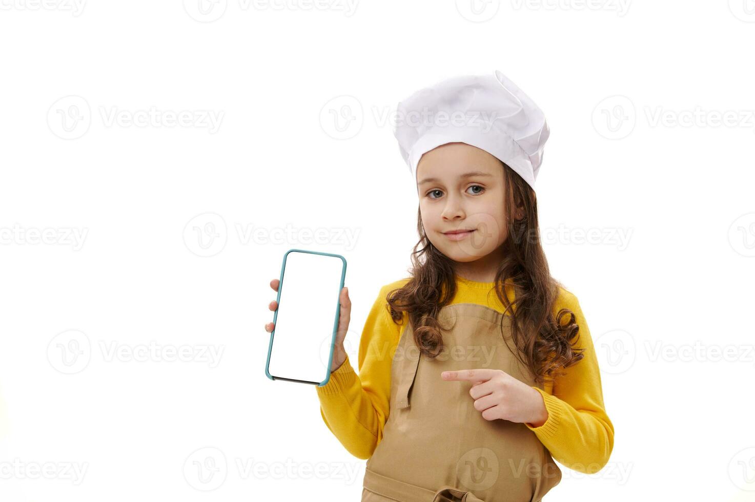 Little chef confectioner in chef's hat and apron, points at white blank screen of smartphone, ad space for mobile apps photo