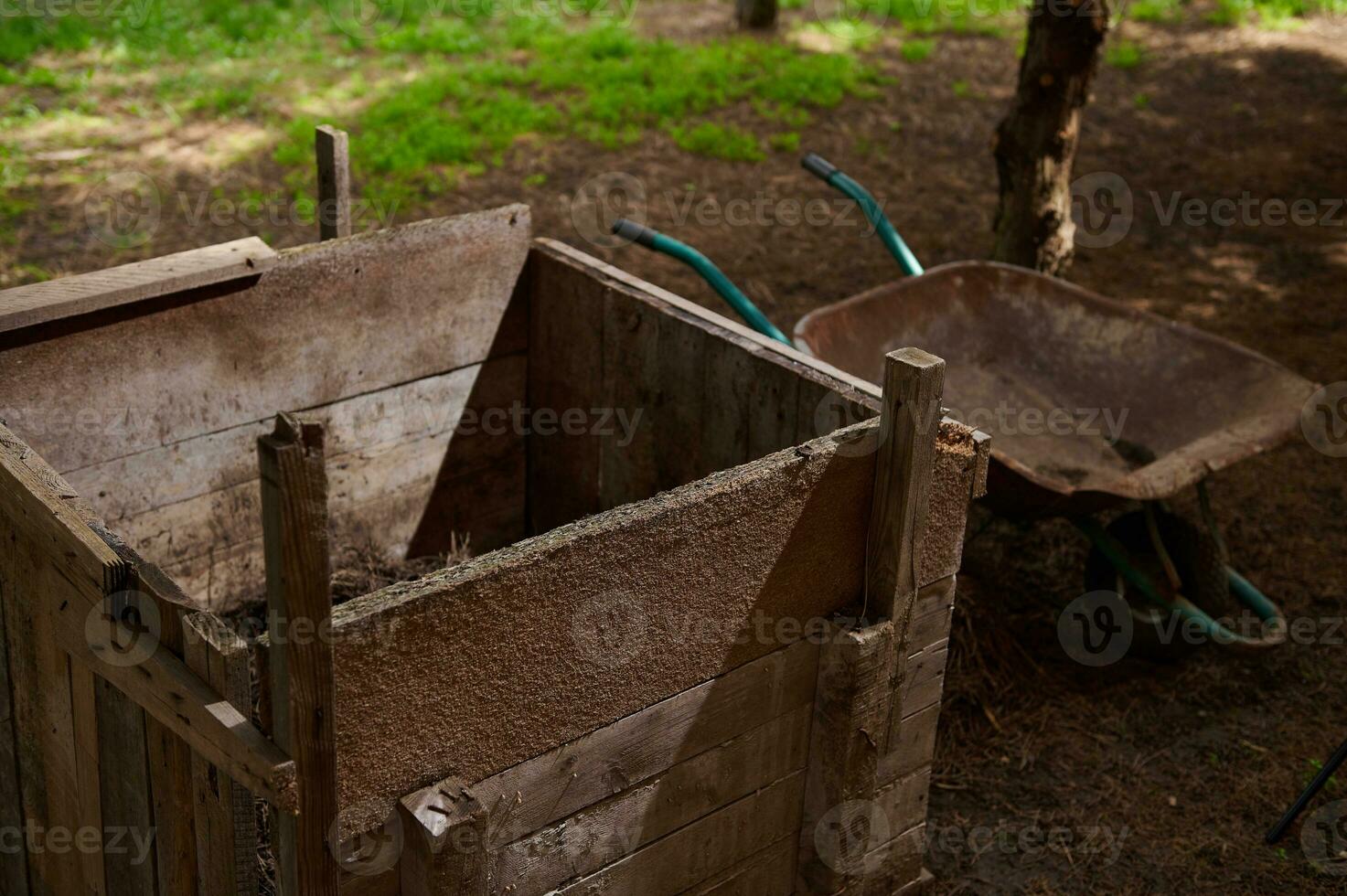 Close-up of wooden compost bin near empty wheelbarrow in the country house backyard. Composting and eco farming concept photo