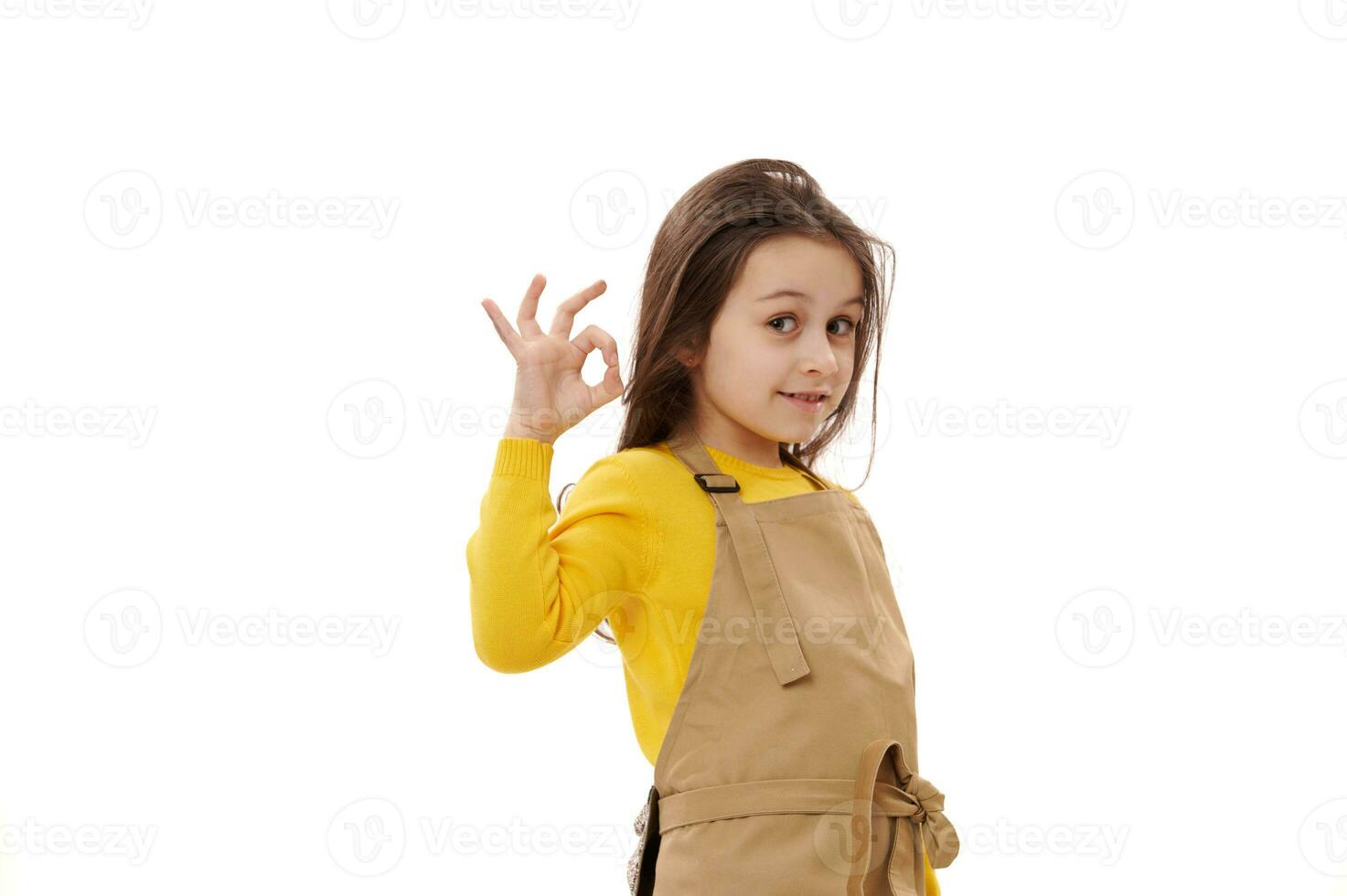 Caucasian kid girl in chef's apron, little waitress in child cafe, gestures with OK sign, expressing positive emotions photo