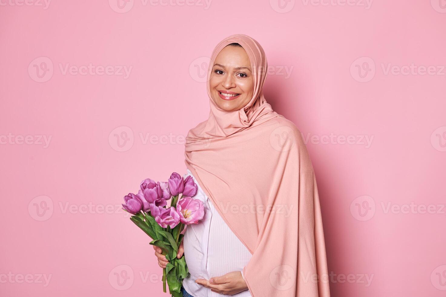 Muslim woman in pink hijab, holding a bouquet of tulips, touching her pregnant tummy, feeling happy by first baby kicks photo