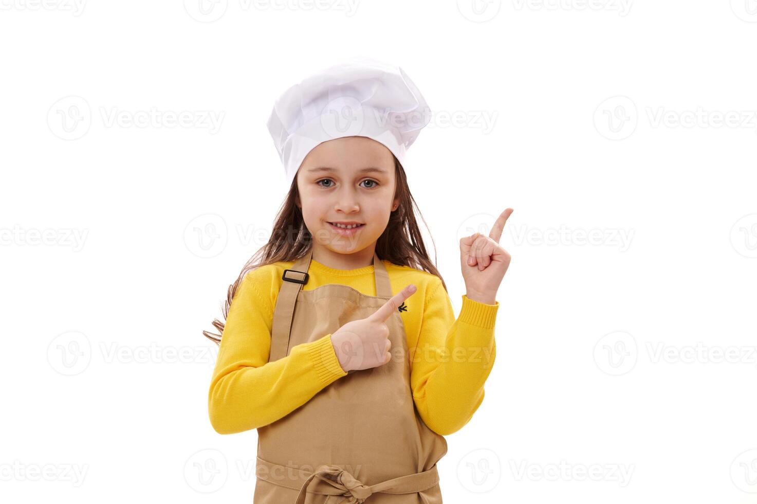 Smiling happy child girl wearing chef's hat and beige kitchen apron, points fingers at copy ad space on white background photo