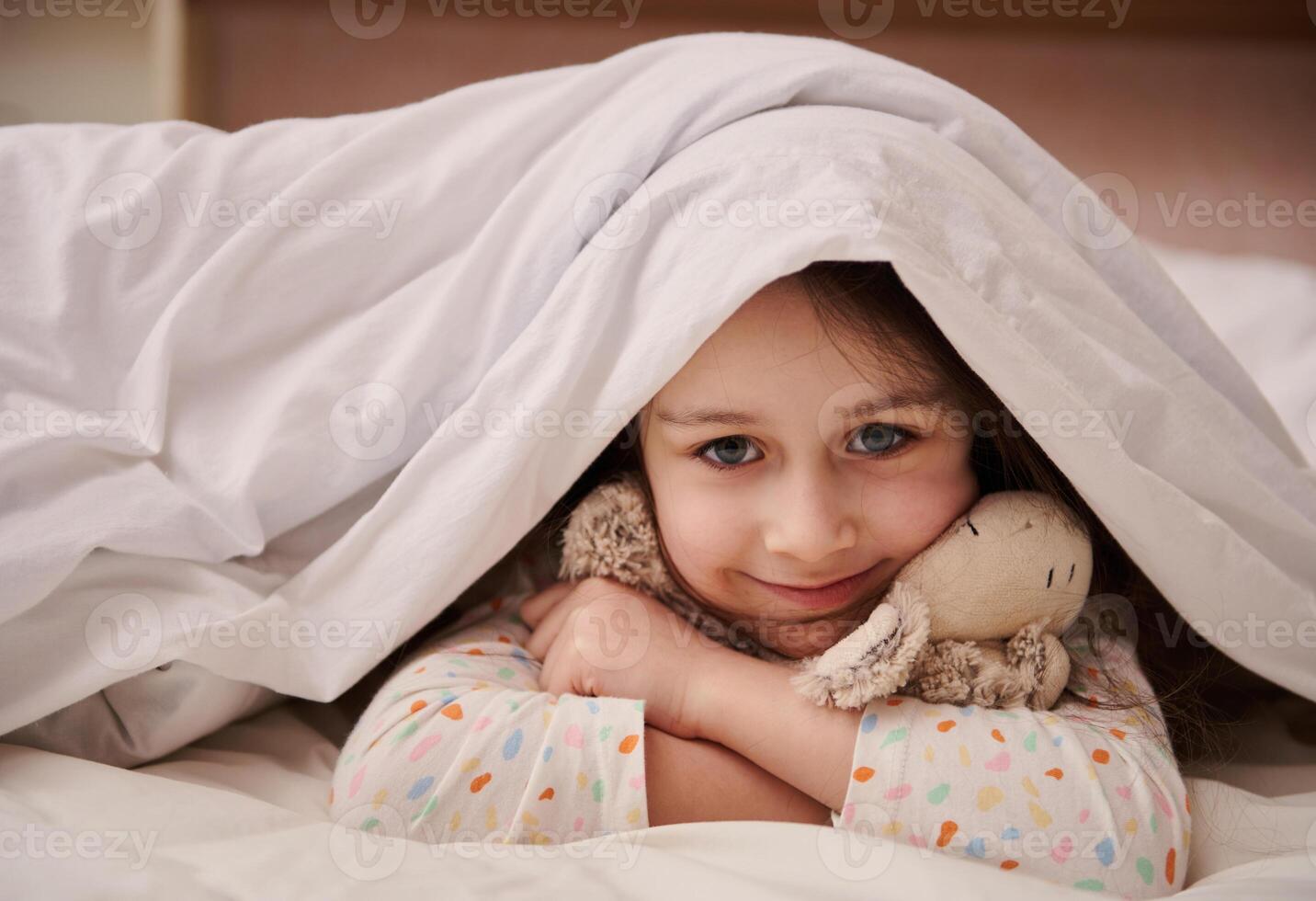 Lovely child girl picking out from soft blanket, cutely smiling looking at camera, gently hugging her cute plush toy photo