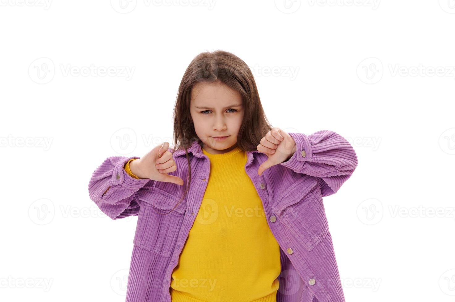 Mischievous little girl gestures with thumbs down, expressing misunderstanding and dissatisfaction, looking at camera photo