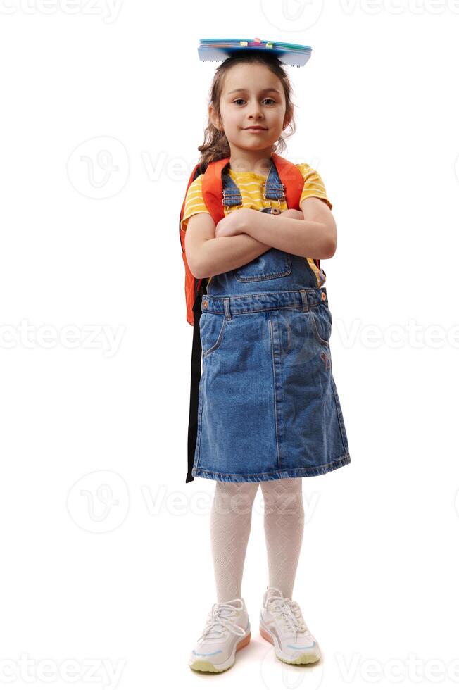 Full size portrait smart schoolgirl with book on her head, smiles at camera, poses with arms crossed on white background photo