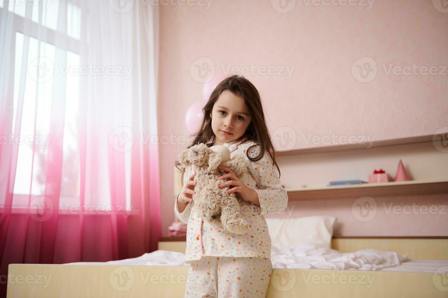 Adorable little child girl in white pajamas, hugging her plush toy sheep and looking at camera in her cozy light bedroom photo