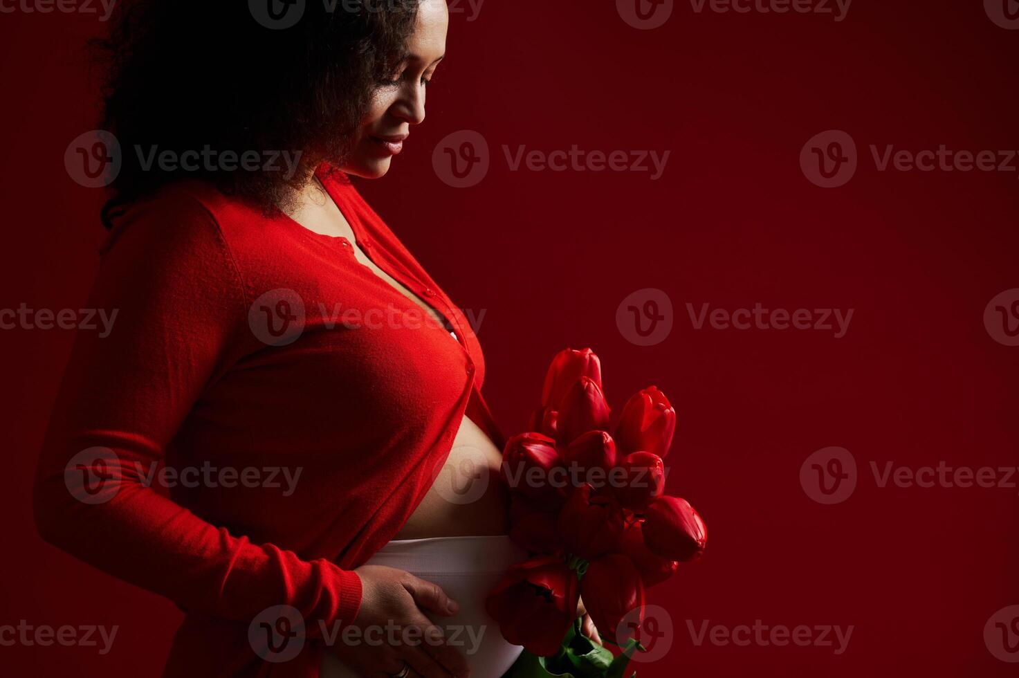 Cropped view of a pregnant woman, expectant mother touching her belly, posing with red tulips, isolated on red backdrop photo